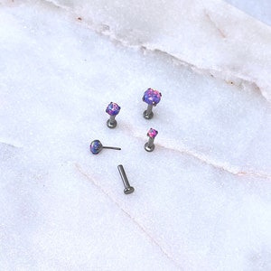 May include: An assortment of small, silver-toned body jewelry pieces with iridescent purple and pink gemstone tops. The jewelry is displayed on a white and gray marble surface. The pieces include labret studs and a post.