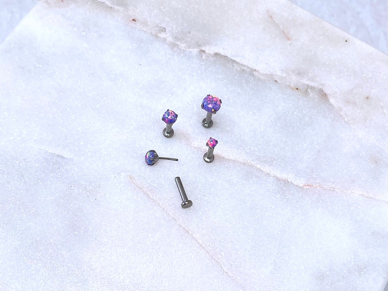 May include: An assortment of small, silver-toned body jewelry pieces with iridescent purple and pink gemstone tops. The jewelry is displayed on a white and gray marble surface. The pieces include labret studs and a post.