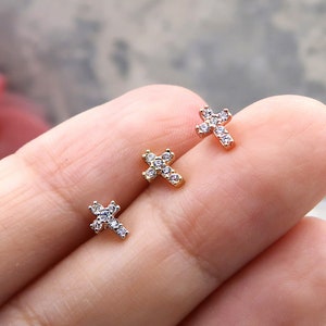 May include: Three small, gold cross earrings with clear cubic zirconia stones. The earrings are on a finger.