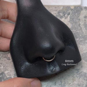 May include: A close-up of a black nose ring on a mannequin. The ring is 8mm in diameter and 16 gauge thickness.