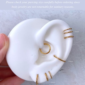 May include: A close-up of a model ear with different sizes of gold hoop earrings. The earrings are 6mm, 8mm, and 10mm in diameter. The text on the image reads: "(*) Size recommendation on this image is just a sample guideline. Each person's ear anatomy is different. Please check your piercing size carefully before ordering since body jewelry are not returnable for sanitary reasons."