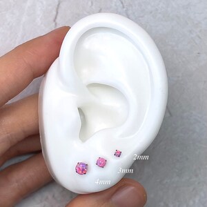 May include: Three pink opal stud earrings in 2mm, 3mm, and 4mm sizes are displayed on a white ear model. The earrings have a round shape and a shimmering, iridescent appearance. The sizes are labeled on the ear model.