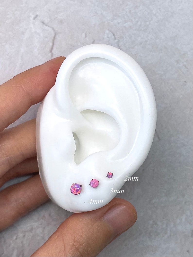 May include: Three pink opal stud earrings in 2mm, 3mm, and 4mm sizes are displayed on a white ear model. The earrings have a round shape and a shimmering, iridescent appearance. The sizes are labeled on the ear model.