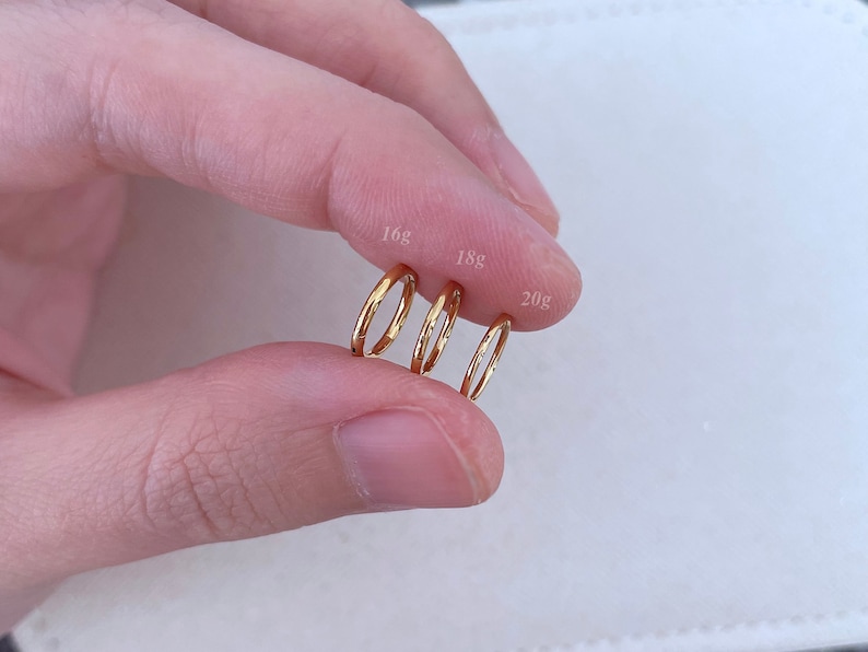 May include: Three gold hoop earrings of different sizes, labeled 16g, 18g, and 20g. The hoops are held in a hand.