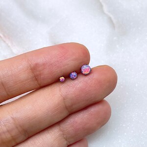 May include: Three small, round opal earrings in varying sizes, displaying a mix of pink, purple, and iridescent colors. The earrings are set on a silver post. The earrings are displayed on a white surface.