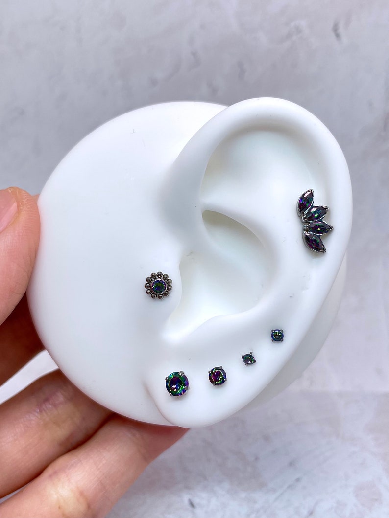 Pode incluir: Um conjunto de cinco pe&ccedil;as de joias para o corpo com pedras preciosas de arco-&iacute;ris iridescentes. As pe&ccedil;as incluem um brinco em forma de flor, um brinco redondo, um brinco quadrado e um grupo de tr&ecirc;s brincos em forma de l&aacute;grima.