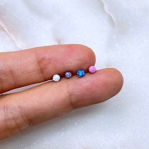 May include: A close-up of five small, round opal stud earrings in a variety of colors, including white, purple, blue, and pink. The earrings are displayed on a person's finger against a white marble background.