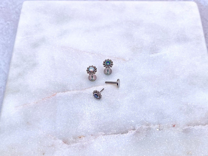 May include: Three silver body jewelry pieces with iridescent gemstones set in a flower design. The jewelry is on a white marble surface.