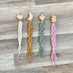 May include: Four handmade macrame baby teethers with wooden animal and geometric shapes. The teethers are in white, mustard yellow, light green, and pink.