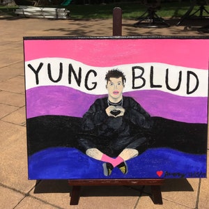 Op de afbeelding: Een schilderij op canvas met de tekst "YUNG BLUD" en een persoon die een hart vormt met zijn handen. De achtergrond is een mix van roze, wit, paars, zwart en blauw. Het schilderij staat op een houten ezel.