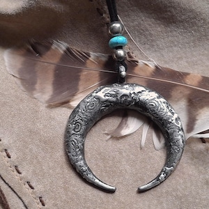 Puede incluir: Un collar con un colgante de media luna de plata con superficie texturizada. El collar presenta cuentas de color azul turquesa y un cordón de cuero marrón.