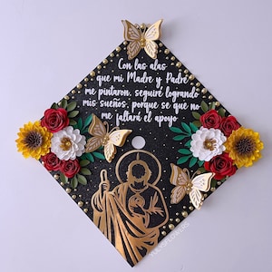 May include: A black graduation cap decorated with gold butterflies, flowers, and the image of a saint. The cap has the text "Con las alas que mi Madre y Padre me pintaron, seguiré logrando mis sueños, porque se que no me faltará el apoyo."