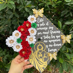 May include: A graduation cap decorated with red, white, and green paper flowers, gold butterflies, and gold glitter. The cap also features a gold silhouette of a religious figure and the text "Este es solo el comienzo, voy por mas! Gracias a todos los que me apoyaron y me dieron fuerza."