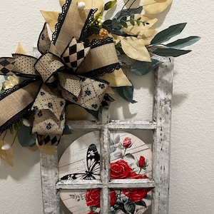 May include: A rustic white window frame with a round, floral-patterned panel featuring a butterfly and red roses. The window frame is decorated with a black and brown bow and a leafy garland.
