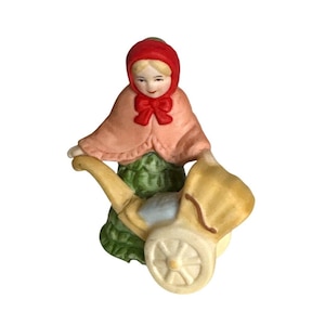 May include: A ceramic figurine of a woman in a red hood and pink cape, pushing a yellow baby carriage. The woman is wearing a green dress. The figurine has a vintage aesthetic.