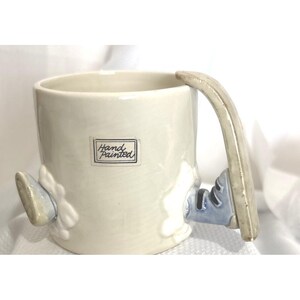 May include: A cream-colored ceramic mug featuring a rectangular label with the text Hand Painted. The mug has a sculpted handle and decorative elements resembling blue and white boots or shoes attached to the sides of the cylindrical body.