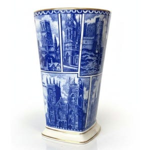 May include: A tapered ceramic vase featuring a blue and white transferware pattern depicting various architectural landmarks and cathedrals. The vessel has a square base with rounded corners and a gold-toned rim along the top edge.