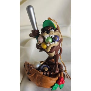 May include: A brown Tasmanian Devil figurine wearing a green baseball cap, holding a silver baseball bat, and biting into a cluster of small, colorful round ornaments. The figure stands inside a brown baseball glove base with red holly berries.