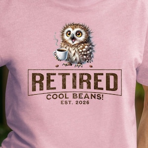 以下が含まれることがあります： コーヒーを飲むフクロウのグラフィックが特徴の薄いピンク色のTシャツ。「RETIRED COOL BEANS! EST. 2026」の文字が下にプリントされています。半袖とクルーネックのシャツです。