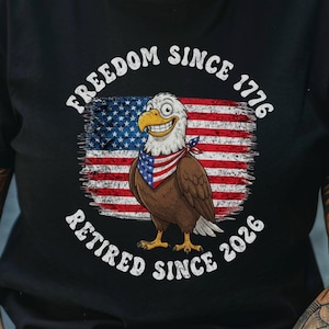 May include: Black t-shirt with a cartoon eagle design set against an American flag. The eagle is wearing a red, white, and blue bandana. The shirt's text reads "FREEDOM SINCE 1776" and "RETIRED SINCE 2026".