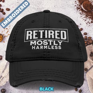 Könnte beinhalten: Schwarze Baseballkappe im Used-Look mit den Worten "RETIRED MOSTLY HARMLESS" in weißen Blockbuchstaben. Das Wort "EMBROIDERED" ist in blauer Schrift auf der oberen linken Seite der Kappe aufgedruckt. Das Wort "BLACK" ist in blauer Schrift aufgedruckt.