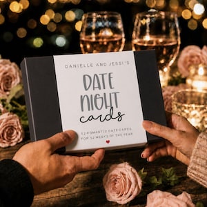 May include: A black rectangular box with a white centre label featuring the text Danielle and Jessi's Date Night Cards, 52 romantic date cards for 52 weeks of the year, and a small red heart icon, held between two hands against a dark background.
