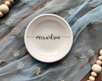 Puede incluir: Un plato pequeño redondo de cerámica color hueso con el texto negro en cursiva "mrs.wilson" centrado en la superficie. El plato está colocado sobre una tela azul claro transparente con estampado floral y cuentas de madera en las esquinas.