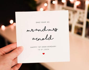 Op de afbeelding: Een witte kaart met zwarte tekst die luidt "ONE YEAR AS mr and mrs arnold HAPPY 1ST ANNIVERSARY 13.01.2024" en een klein rood hart. De kaart wordt in een hand gehouden.