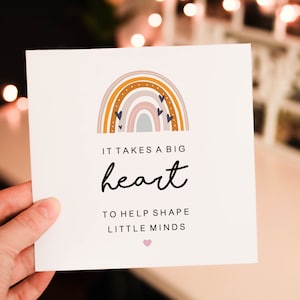May include: A white card with a rainbow illustration and the text "IT TAKES A BIG heart TO HELP SHAPE LITTLE MINDS." The rainbow features shades of brown, pink, and blue. A small pink heart is at the bottom.