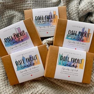 May include: Five brown cardboard boxes with white labels reading "DATE NIGHT cards" featuring city skylines. Each box is labelled with a different city: Adelaide, Brisbane, Sydney, Melbourne, and Perth. The boxes are on a grey, textured surface.