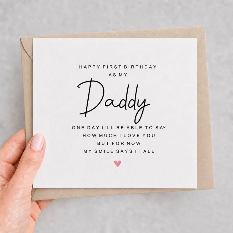 Op de afbeelding: Een vierkante witte wenskaart met zwarte tekst: HAPPY FIRST BIRTHDAY AS MY Daddy ONE DAY I'LL BE ABLE TO SAY HOW MUCH I LOVE YOU BUT FOR NOW MY SMILE SAYS IT ALL. Een klein roze hartje staat onder de tekst op een bruine envelop.