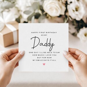 Op de afbeelding: Een vierkante witte wenskaart met zwarte tekst die luidt: HAPPY FIRST BIRTHDAY AS MY Daddy ONE DAY I'LL BE ABLE TO SAY HOW MUCH I LOVE YOU BUT FOR NOW MY SMILE SAYS IT ALL. Een klein roze hartje is onder de tekst op de kaart gedrukt.