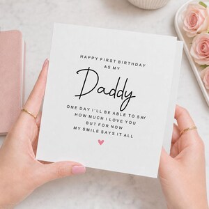 Op de afbeelding: Een vierkante witte wenskaart met zwarte tekst die luidt: HAPPY FIRST BIRTHDAY AS MY Daddy ONE DAY I'LL BE ABLE TO SAY HOW MUCH I LOVE YOU BUT FOR NOW MY SMILE SAYS IT ALL. Een klein roze hartje is onder de tekst gedrukt.
