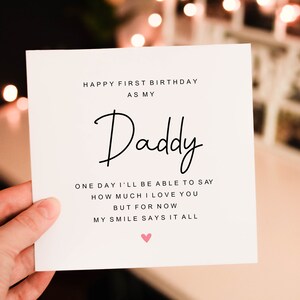Op de afbeelding: Een vierkante witte wenskaart met zwarte tekst die luidt: HAPPY FIRST BIRTHDAY AS MY Daddy ONE DAY I'LL BE ABLE TO SAY HOW MUCH I LOVE YOU BUT FOR NOW MY SMILE SAYS IT ALL. Een klein roze harticoontje is onder de tekst gedrukt.