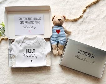 Puede incluir: Un body de bebé blanco con el texto Hello Daddy y un pequeño corazón, dentro de una caja de regalo con una tarjeta que dice Only the best husband gets promoted to be Daddy, junto a un oso de peluche con peto azul a rayas y una tapa con el texto To the best husband.