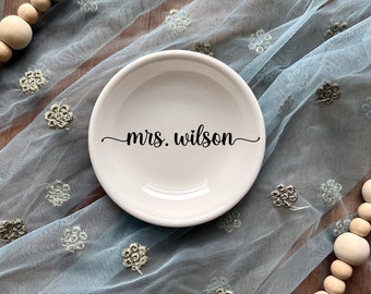 Puede incluir: Un plato pequeño redondo de cerámica color hueso con el texto negro "mrs. wilson" impreso en el centro. El plato está colocado sobre una tela azul claro transparente con estampado floral, acompañado de un collar de cuentas de madera natural.