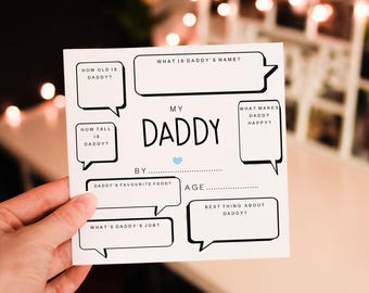 Puede incluir: Una tarjeta blanca para rellenar con el texto "MY DADDY" y preguntas como "¿Qué edad tiene papá?" y "¿Qué hace feliz a papá?" Un pequeño corazón azul está debajo del título. La tarjeta está sujeta por una mano.