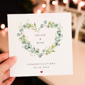 May include: A white card with a green eucalyptus wreath in the shape of a heart. The text on the card reads "Helen & Mike Congratulations 05.04.2024" with a small red heart at the bottom.
