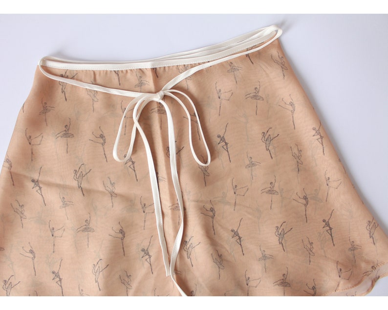 Pu&ograve; includere: Una gonna avvolgente beige con un bordo bianco e una stampa di ballerine. La gonna si lega in vita con un nastro bianco.