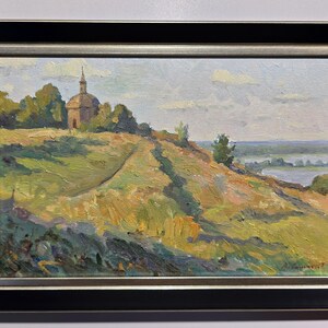May include: Oil painting of a landscape with a small church on a hill. The artwork features a path leading up to the church, with a river and sky in the background. The painting is framed in a black and silver frame.