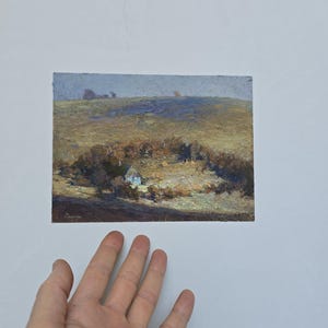 May include: An oil painting on a rectangular canvas depicting a landscape with a small house nestled in a cluster of trees. The scene features a textured, earthy color palette of browns, ochres, and muted blues under a soft, overcast sky.