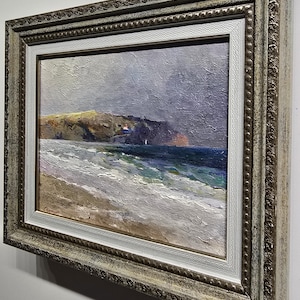 May include: Oil painting of a coastal landscape with a beach, ocean, and cliffs under a cloudy sky. The artwork is framed in a decorative, ornate, gold-toned frame with a white inner border. The painting depicts a serene seascape.