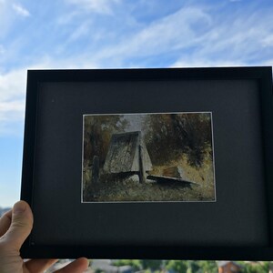 May include: A framed landscape painting with a black frame. The artwork depicts a rustic structure in a forest setting, with muted tones of brown, green, and gray. The background shows a blue sky with clouds.