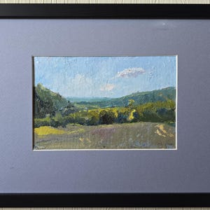May include: Oil painting landscape framed in black. The artwork depicts a valley with green trees and a field of yellow and purple flowers. The sky is blue with a few white clouds. The painting is set in a gray mat.