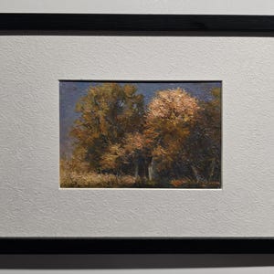 May include: Framed landscape painting featuring a vibrant autumn scene. The artwork depicts trees with golden and reddish-brown foliage under a blue sky. The painting is set within a white mat and a black frame, creating a classic presentation.