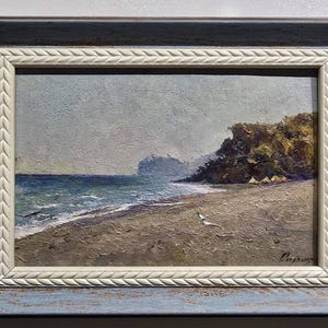 May include: Oil painting of a coastal scene with a beach, ocean, and cliffs under a cloudy sky. The artwork is framed in a blue and white decorative frame. The painting depicts a serene beach landscape.