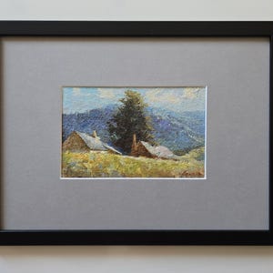 May include: Framed landscape painting depicting a rural scene with two small houses and a tall tree in the foreground. The background features rolling blue mountains under a light blue sky. The artwork is framed in a black frame with a gray mat.
