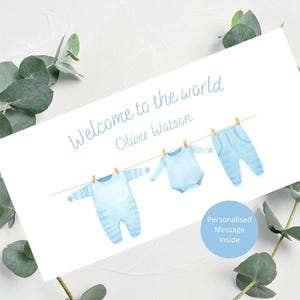 May include: A greeting card with the text "Welcome to the world" and a baby's name. The card features a watercolor illustration of baby clothes hanging on a clothesline. A blue circle indicates a personalized message.