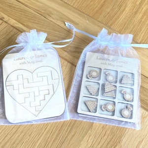 K&ouml;nnte beinhalten: Zwei wei&szlig;e Organza-Beutel mit Kordelzug und Holzspielen. Ein Beutel enth&auml;lt ein herzf&ouml;rmiges Puzzle, der andere ein Tic-Tac-Toe-Spiel mit Ring- und Torten-Spielsteinen. Graviert mit "Lauren & James 10th May 2026".