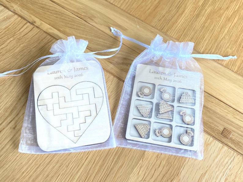 K&ouml;nnte beinhalten: Zwei wei&szlig;e Organza-Beutel mit Kordelzug und Holzspielen. Ein Beutel enth&auml;lt ein herzf&ouml;rmiges Puzzle, der andere ein Tic-Tac-Toe-Spiel mit Ring- und Torten-Spielsteinen. Graviert mit "Lauren & James 10th May 2026".